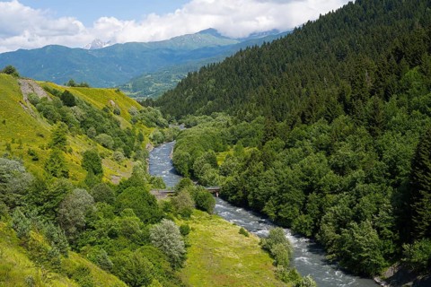 River flows through green mountains and forests with a peaceful summer atmosphere under cloudy