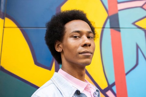 A non-binary person with an afro hairstyle gazes confidently at the camera, standing against a