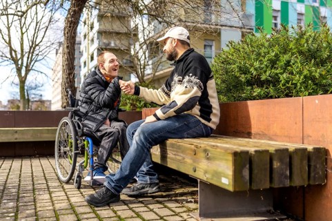 Two friends shaking hands while one sits in a wheelchair, both enjoying a sunny day together in the