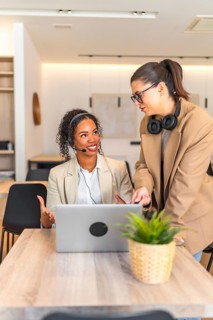 Two female customer service representatives are collaborating on a laptop, discussing work related