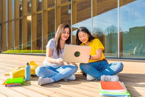 Frontal photo of two multi-ethnic female friends using laptop sitting on the university campus