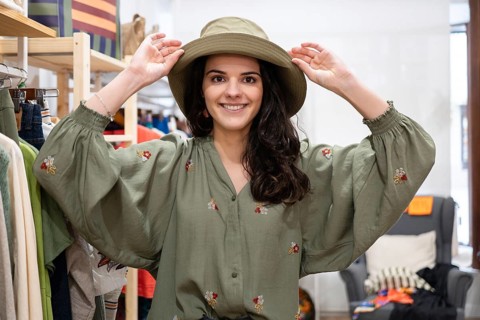 A woman smiles in a clothing store, wearing a green hat and embroidered dress. The setting is