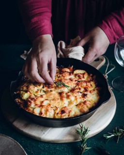 A close up of hands garnishing a cheesy potato gratin with fresh rosemary in a cast iron skillet.