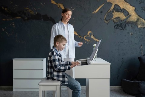 A young boy in a checkered shirt focuses on playing the piano in a modern room. His teacher,