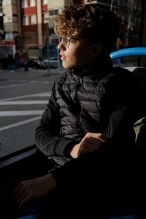 A young man with glasses sits on a bus, looking thoughtfully out the window. Sunlight highlights