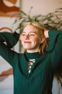 A teenager with a bright smile poses indoors, arms raised with confidence. The warm ambiance and