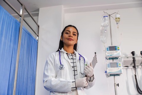 A confident female doctor stands in a hospital setting, holding a clipboard and wearing a white