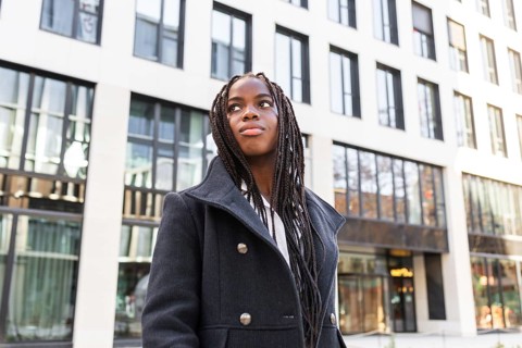 A young African American businesswoman with braids, wearing a stylish coat, takes a break outdoors