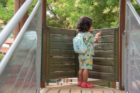 Back view of a young child in colorful attire and backpack explores a wooden playground bridge.
