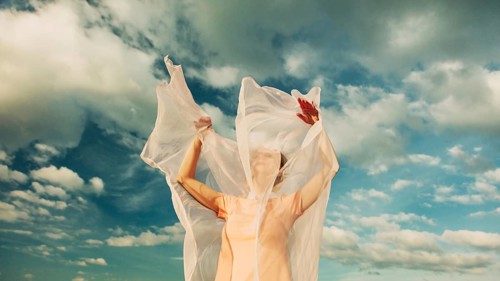 A woman stands with arms raised, her face partially obscured by translucent fabric flowing in the