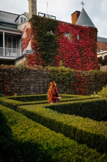 A woman with long red hair explores a lush green hedge maze in an urban garden in Montreal. The