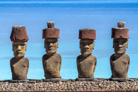 Four Moai statues with red Pukao headpieces on a stone platform in front of a turquoise Pacific