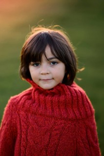 A young girl wearing a vibrant red sweater poses outdoors, her expression thoughtful. The setting
