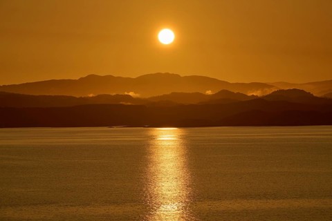 A soft orange sunrise behind the dark silhouettes of mountains and a calm sea, sunbeams reflecting
