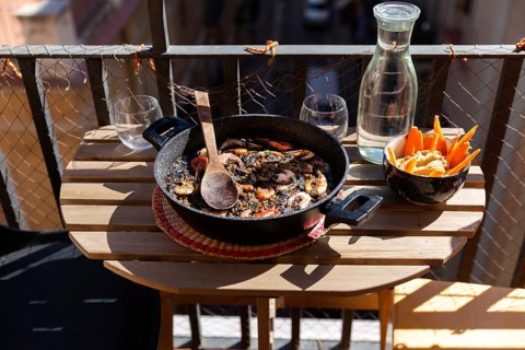 A rustic table on a balcony in Barcelona holds a delicious black paella with seafood dish, water,