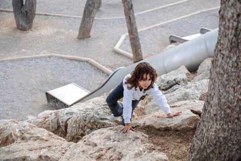 A young girl explores a rocky terrain in a park surrounded by trees. The afternoon light casts a