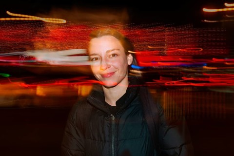 A smiling woman in a black jacket stands against blurred, colorful light trails in an street night