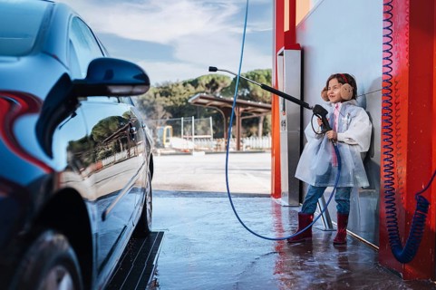 A young girl at a car wash uses a high pressure washer. She smiles, wearing earmuffs and boots,