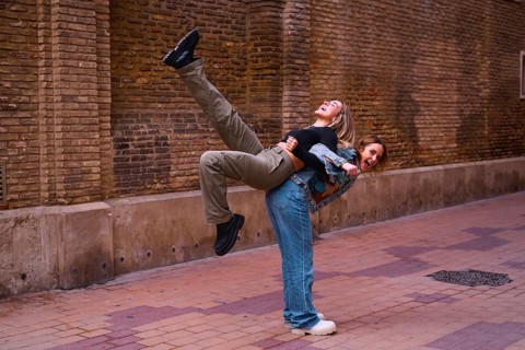 Two young woman friends share a playful moment on a cobblestone street. One joyfully lifts the