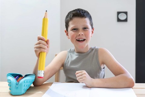 Smiling child in a classroom holding an oversized pencil, symbolizing creativity and learning. A