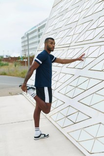 A black male athlete stretches his leg against a sleek, geometric wall. He wears sportswear,