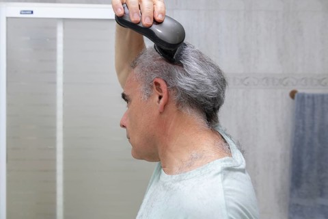 A mature man is seen trimming his hair with an electric trimmer in a tiled bathroom. The image
