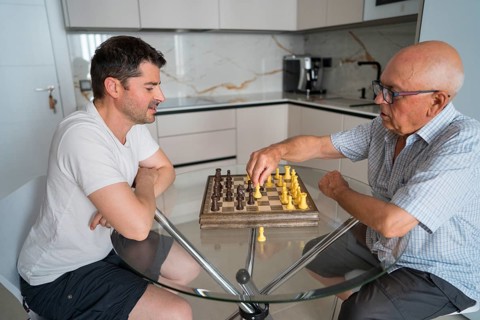 A father and adult son engage in a friendly game of chess at home, strengthening their bond. The