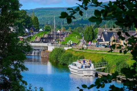 A boat passes a lock in a village with green surroundings and hills, Caledonian Canal, Fort