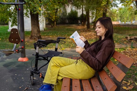A woman with cerebral palsy sits on a park bench, reading a pamphlet. A walker is parked beside