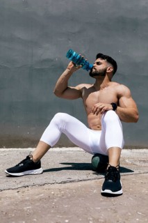 Athletic man wearing white leggings and running shoes drinks water while seated outdoors against a