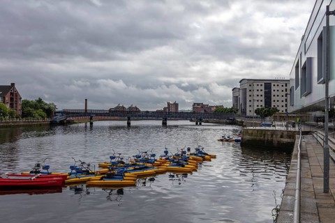 River with anchored boats, surrounded by buildings and a bridge under a cloudy sky, Belfast