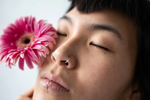 Close-up portrait of an Asian woman with closed eyes. She holds a pink flower near her face,
