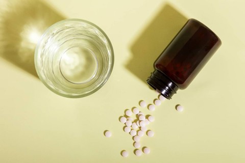 Overhead view of white pills scattered from a brown bottle next to a clear glass on a light