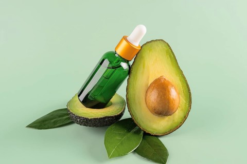 A glass dropper bottle of avocado oil elegantly balanced on half an avocado with green leaves and a