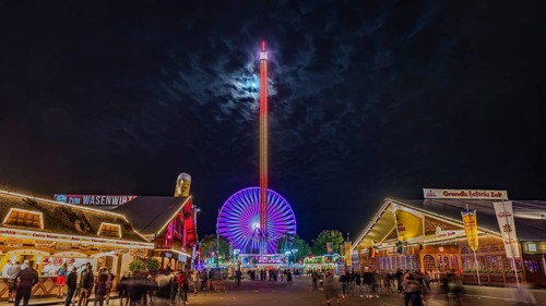 A brightly lit Ferris wheel and marquees at night at a lively funfair with numerous visitors,