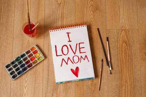 A heartfelt I LOVE MOM message is painted in red on a spiral notebook, surrounded by brushes,