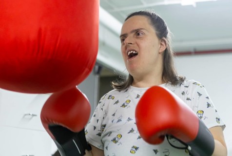 A woman enthusiastically punches a red boxing bag at a mental health residence, using boxing as a