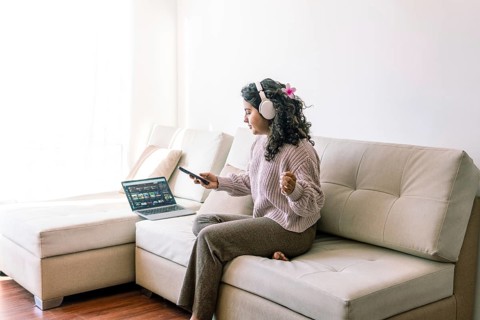 A young latin woman enjoying music with headphones, sitting on a comfy sofa A laptop is open on the