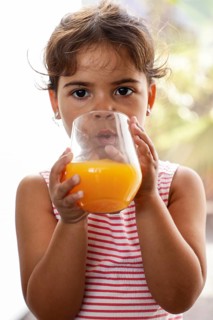 A young girl sips fresh orange juice from a glass. She wears a pink and white striped tank top,