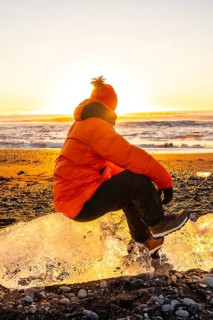 A man in an orange jacket sits on a rock near the ocean. The sun is setting, casting a warm glow