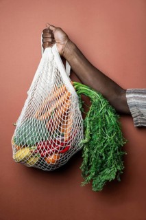 A person holds a reusable mesh bag filled with fresh vegetables, including carrots and peppers,