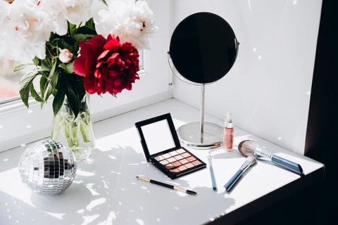 Vibrant red and white peonies in a clear glass vase beside a makeup vanity with a mirror, makeup