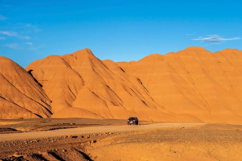 A vehicle traverses a winding road amidst the striking orange sandy mountains under a clear blue