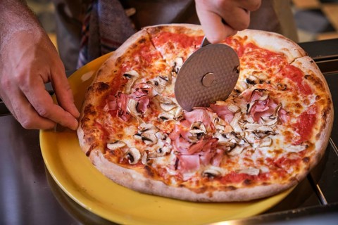 Cook cutting freshly baked pizza with ham and mushrooms using a round pizza cutter