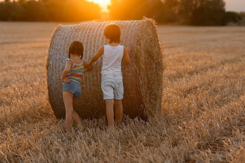 Back view of two unrecognizable children, a boy and a girl, play around a large hay bale in a field