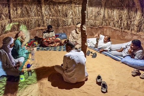A group of people sit in a traditional hut with a sandy floor and relax in conversation, A group of