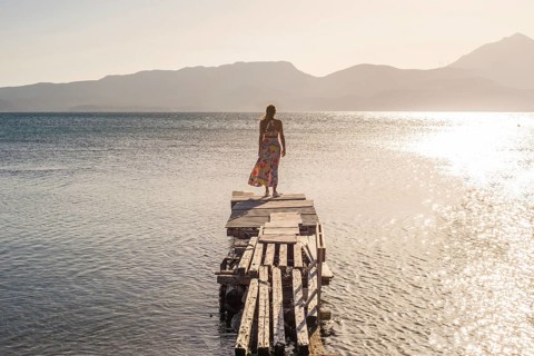 A woman stands on a rustic pier at Papikinou Beach, Milos Island, Greece, gazing at the tranquil
