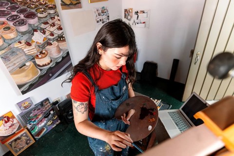Young Latin woman focused on painting a canvas in her home art studio. Surrounded by paintings, she