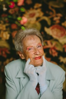 An elegant elderly woman in a light jacket and white gloves poses joyfully with a floral backdrop.