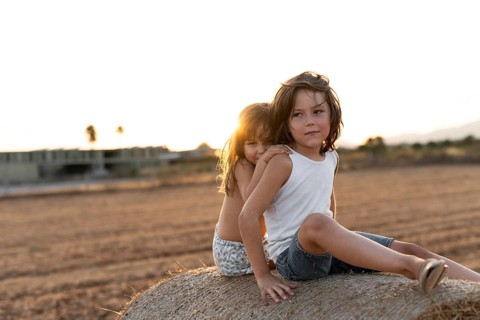 Two children sit together on a hay bale, enjoying the warm sunshine in a rural setting. The